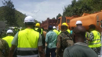 A group of people are gathered around a large, orange dump truck in Sake, Democratic Republic of Congo. The truck is parked on a dirt road with a hill in the background. The people are wearing a variety of clothing, including some who are wearing yellow safety vests. Many of the people are wearing hard hats. The people in the foreground are looking towards the truck, while others are looking around. Several people are holding onto a large backpack. Some people are talking to each other. The dump truck is partially obscured by trees in the background. The road is dirt and dusty. The weather is sunny and clear.