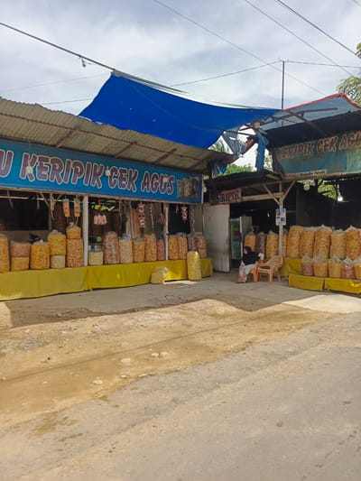 The image depicts an outdoor food stall in Sawang, Indonesia, specializing in "keripik" (chips). The stall features a long display counter covered with yellow fabric, showcasing numerous large plastic bags filled with various types of chips. Behind the counter, a blue tarp and corrugated metal sheeting provide shelter for the stall. A sign with blue lettering reads "U KERIPIK CEK AGUS," indicating the name of the establishment.
In the middle section of the stall, a woman wearing a black headscarf and a dark-colored dress is seated on a plastic chair, facing towards the right of the image. Two other plastic chairs are visible nearby. A refrigerator is also present behind the counter. The ground in front of the stall is a mix of dirt and asphalt, with some debris scattered around. The overall scene suggests a typical roadside vendor in a tropical environment.