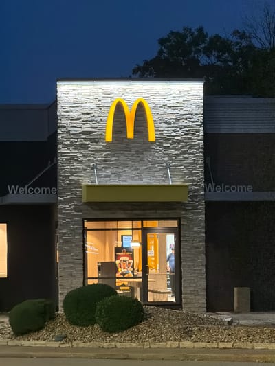 The image shows the exterior of a McDonald's restaurant at night. The building is constructed with a stone facade and features the iconic golden arches logo illuminated at the top. A yellow overhang is positioned above the entrance. The entrance itself is a glass door leading into the well-lit interior of the restaurant, where a worker in uniform can be seen behind the counter. A promotional poster for the FIFA World Cup is visible inside. The foreground is landscaped with gravel and several rounded green bushes. The words "Welcome" are visible on signage to the left and right of the main entrance. The sky above is dark blue, indicating it is nighttime. The location is identified as Maryland Heights, United States of America.