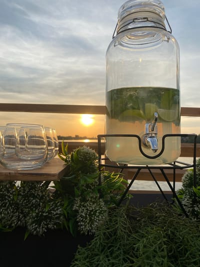 Here's a detailed description of the image:
**Scene:** The photograph appears to have been taken outdoors, possibly on a deck or patio, with a sunset visible in the background. The lighting suggests it is either early morning or late afternoon/early evening. The location is likely not in the city of Aramil, Russia, as there's no supporting evidence in the image to suggest that particular location.
**Objects:**
* A large glass beverage dispenser filled with a light-colored liquid, possibly lemonade or water with lime slices. It has a spigot for dispensing the liquid.
* The dispenser sits on a black metal stand.
* Next to the dispenser are two clear drinking glasses stacked together.
* Decorative greenery, including leafy plants with small white flowers and trailing, vine-like plants, is arranged around the dispenser and glasses.
* A wooden structure, possibly a fence or wall, forms a backdrop behind the setup.
**Environment:**
* The sky is visible in the background, with the sun setting or rising, creating a warm, golden glow.
* There is land or vegetation visible on the horizon.
* The overall mood is serene and relaxed.
**Actions/Interactions:** No people or active interactions are visible. The scene is a still life arrangement.
**People:** No people are visible in the image.