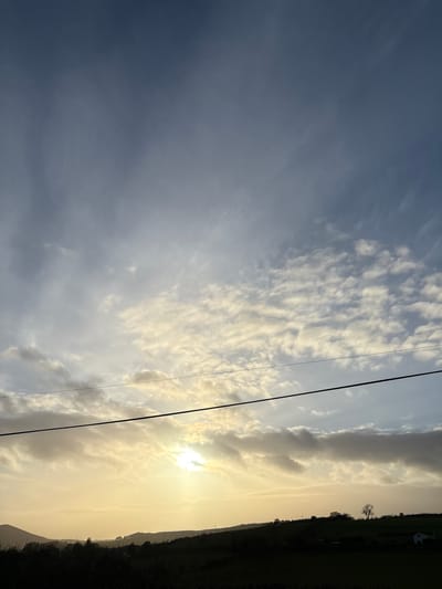The image depicts a rural landscape at either sunrise or sunset, located near the city of Stiperstones, United Kingdom. The sky occupies approximately the upper two-thirds of the frame, transitioning from a dark blue-grey at the top to a bright yellow-orange and white glow towards the lower-middle where the sun is partially obscured by clouds. Clouds, ranging from scattered altocumulus to more diffuse stratus formations, are present across the sky, particularly concentrated around the sun. A prominent dark utility wire or cable traverses horizontally across the mid-section of the image, positioned above the silhouetted horizon line. The lower third of the image features a dark, silhouetted hilly landscape. Multiple undulating hills and ridges are visible, sparsely populated with trees, some appearing bare. Faint silhouettes of utility poles and at least one structure, possibly a house with a lighter-colored wall or roof, are discernible on the distant hills. The foreground consists of dark, indistinct vegetation, likely fields or shrubbery. No people, animals, or explicit actions are visible within the frame.