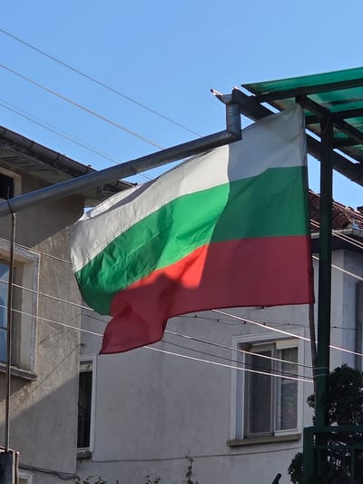 The image shows a Bulgarian flag hanging from a structure attached to a building. The flag is a tricolor, with white, green, and red stripes. The building is light gray and has windows. Power lines are visible in the background against a blue sky. The location is identified as Kaleytsa, Bulgaria.