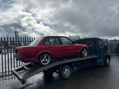 A flatbed truck is transporting a red car and another vehicle in Shotts, United Kingdom. The truck is parked next to a metal fence. The sky is overcast with dark clouds. A communications tower is visible in the background behind the fence. The ground appears to be wet.