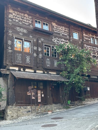 The image shows the facade of a historic wooden building in Grabs, Switzerland. The building is adorned with white painted ornamental script and decorative patterns. Multiple windows with white frames are visible, some with checkered panes. A large rose bush with pink flowers is growing up the side of the building, partially obscuring some of the windows and decorative elements. Below the upper floors, there is a covered entrance with a wooden roof supported by beams. The entrance area features wooden doors and windows, with several framed pictures and metal sculptures displayed on a stone wall to the left. The building is situated on a cobblestone street that slopes upwards. The sky is overcast.