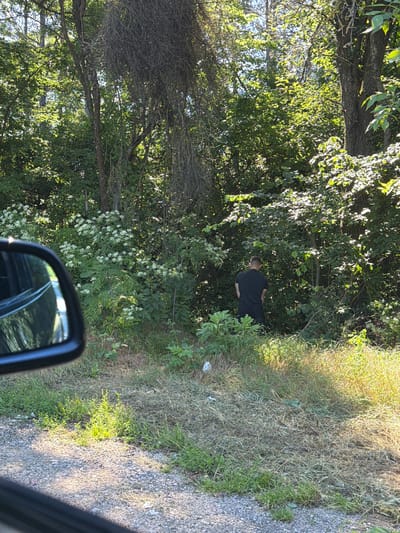 The image shows a man in a black t-shirt and black shorts urinating in a wooded area. He is standing with his back to the camera, facing the dense foliage. The environment is a natural setting with trees, bushes, and dry grass. There is also a car's side mirror visible in the foreground, reflecting the surroundings. The location is identified as the city of Garnia, Bulgaria.