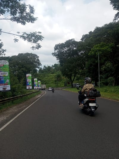 The image depicts a paved road in Niz Lakuk, India, under an overcast sky. In the foreground, the rear of a white scooter, bearing license plate "B 5398 BZL," travels away from the viewer. A person wearing a striped helmet, light-colored long-sleeved shirt, and dark pants operates the scooter, with a passenger in dark attire seated behind. A black bag is secured to the back of the scooter. The road curves slightly to the right. To the left, a metal guardrail separates the road from a grassy verge and several vertical billboards displaying advertisements, including text "IDUL FITRI 1447 H," "JUARA," and "KRETEK KLK." Further down the road, another motorcycle and a white truck are visible, also heading away. The immediate environment is characterized by dense green vegetation, large trees, and rising hills covered in foliage, particularly on the right side of the road where a street lamp post is also present. The background shows more lush green hills beneath a sky filled with grey clouds.