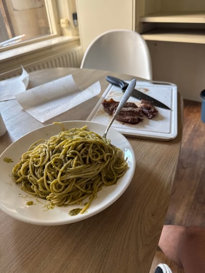 The image depicts a close-up, slightly elevated view of a wooden dining table where a meal is being prepared or consumed. In the foreground, a white circular plate is filled with spaghetti tossed in a green sauce, likely pesto. A silver fork is partially submerged in the pasta, suggesting it is being eaten. To the right of the plate, on a white cutting board, are several slices of cooked meat, possibly steak, alongside a black-handled knife. White paper towels are folded to the left of the plate. In the background, a white chair is visible, and beyond that, a window with blinds and a radiator. A light-colored shelf unit is also present. The overall scene suggests a casual meal setting, possibly within a domestic environment in Deventer, Netherlands, given the hints. The lighting appears to be natural, coming from the window. No specific Deventer landmarks are visible.