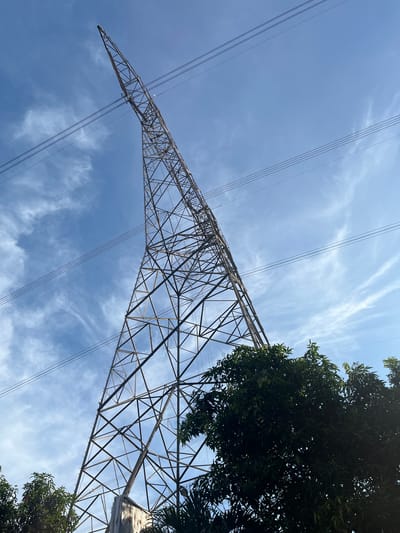 The image shows a low-angle view of a metal electricity pylon against a sky with wispy clouds. The pylon is constructed of a lattice of steel beams, tapering as it extends upwards. Power lines run diagonally across the frame. Tree foliage is visible on the right side of the image. The location is Sukabakti, Indonesia.
