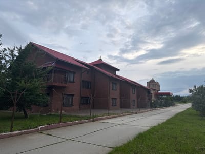 The image shows a building with a red tiled roof, constructed of dark red brick, in Petrovo, Russia. A concrete pathway runs alongside the building, bordered by grass and a chain-link fence. Trees and bushes are visible around the property. In the background, another building with a dome-shaped roof and an ornate balcony can be seen. The sky is cloudy.