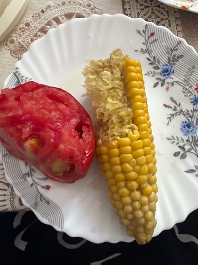 A partially eaten cob of corn and a segment of a tomato are placed on a white plate with a scalloped edge. The plate features a floral pattern with blue and red flowers and grey line accents. The food is presented on a table covered with a patterned tablecloth. The setting appears to be a domestic environment in Taganrog, Russia.