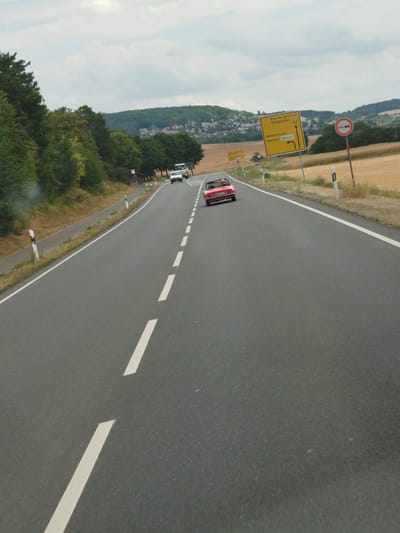 In Lauda, Germany, a red convertible drives down a two-lane asphalt road. The car is in the right lane, heading away from the camera. The road has white dashed lines dividing the lanes. On the right side of the road, there is a yellow sign with directions to Bad Mergentheim, Königshofen, Messelhausen, Marbach, and Weinort. A circular sign with two red cars in silhouette is also visible on the right. The landscape is rural, with trees and fields on either side of the road, and a town on a hill in the background. The sky is overcast.