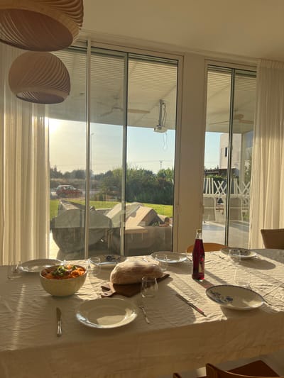A laid table occupies the foreground, draped with a white tablecloth. Plates, cutlery, glasses, a bottle of red liquid, a bowl of food, and a loaf of bread suggest a meal setting. Sunlight streams in through large windows, illuminating the scene. Outside, a veranda and glimpses of a garden with trees and lawn can be seen. Decorative hanging lamps are partially visible on the left.