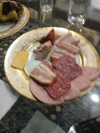 A plate of sliced meats and cheese is arranged on a decorative plate with gold trim. The plate contains slices of salami, ham, bacon, and a piece of cheese. A piece of cooked meat is also visible. The plate is set on a glass table, and in the background, glasses of liquid and another plate of food are partially visible. The scene appears to be an indoor setting, possibly a meal or a gathering. The lighting suggests it could be evening. There is no visible text in the image.