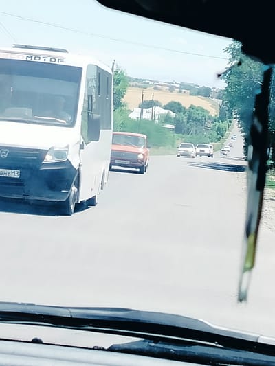The image depicts a view from inside a vehicle, looking out at a road scene. In the foreground, the edge of the vehicle's windshield and wiper are visible. Ahead, a white bus with "MOTOR" written on its side is the most prominent object, followed by an orange car. Several other white cars are visible further down the road, which appears to be a two-lane asphalt road with a gravel shoulder. The surrounding environment is rural, with fields and trees lining the road. The sky is bright and appears to be a clear day with some haze. There are utility poles and wires visible in the distance. The license plate on the white bus reads "BHY 13". The scene is consistent with a road in a settlement like Көлкент, Kazakhstan, likely a highway or main road within or leading out of the town, characterized by its infrastructure and surrounding landscape.