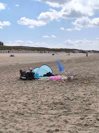 This is a wide shot of a beach under a blue sky with white clouds. In the background are dunes and a line of structures. In the midground, from left to right, are groups of people at a distance and, closer to the viewer, a tent, a wagon, and a collection of beach gear that includes a beach umbrella, a pink inflatable ring, and sand toys. The foreground is sand. The location is a beach near Bergen, Netherlands.