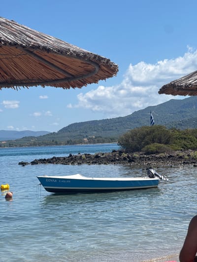 The image depicts a serene coastal scene in Lichada, Greece. Two thatched sun umbrellas partially frame the view of a calm, blue sea. In the foreground, a small blue and white motorboat is moored. The boat has the Greek inscription "ΣΟΥΛΑ Σ. ΑΙΔ. 375" on its side. Two buoys, one yellow and one reddish-orange, float near the boat, connected by a rope.

The middle ground features a rocky outcrop extending into the sea. Beyond this, a lush green hillside dotted with trees rises, forming the backdrop. A Greek flag waves from the top of a bushy area on the hillside. The sky is a vibrant blue with scattered white clouds, suggesting a clear, sunny day. The overall impression is one of tranquility and natural beauty in a Mediterranean setting.