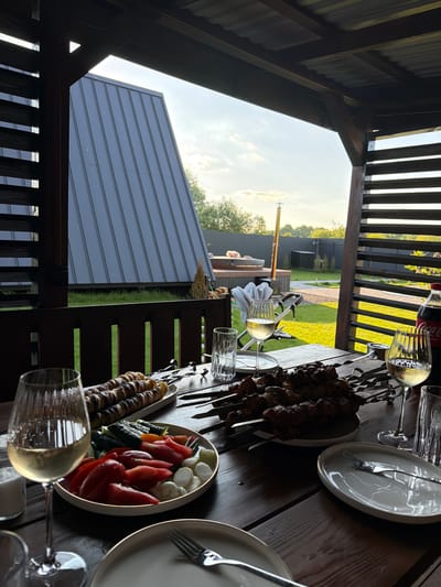 The image depicts an outdoor dining scene at a wooden table under a covered patio. On the table, there are two plates of skewered meat, a platter of sliced vegetables including tomatoes and cucumbers, and two glasses of white wine. A bottle of Coca-Cola and a salt shaker are also visible. In the background, a grassy yard with a fence and a building with a dark gray, sloped roof can be seen. A hot tub is also present in the yard. The setting sun casts a warm light on the scene, indicating it is likely late afternoon or early evening. The overall impression is one of a relaxed meal being enjoyed outdoors. There are no people clearly visible in the scene. The location is identified as Sharovechka, Ukraine.