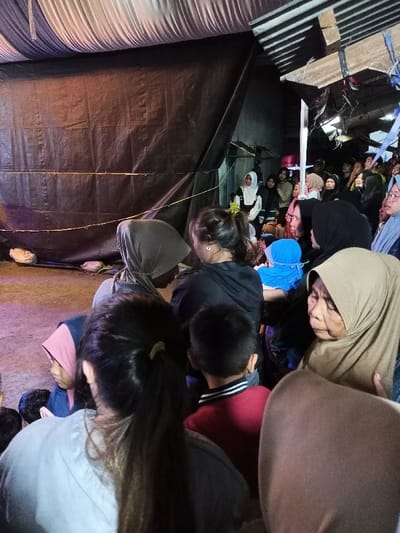 This image captures a crowded community gathering within a dimly lit, temporary structure in Pangalengan, Indonesia. The walls are constructed from large black tarpaulins, with a horizontal string or rope stretched across one. The ceiling appears to be made of draped white and dark fabric, and corrugated metal sheets are visible in the upper right, suggesting a makeshift roof. The ground is unpaved and rough.
A diverse group of people, predominantly women wearing hijabs of various colors (light brown, black, gray, pink), and some children, are gathered closely. In the foreground right, an elderly woman in a light brown hijab looks attentively towards the right. A young boy in a red shirt with a striped collar is seen from behind in the mid-ground. Other individuals include a woman with dark hair tied in a ponytail in the foreground left, seen from the back, and several women in black hijabs. A young girl in a pink hijab is also visible.
The people are looking in various directions, suggesting they are observing an event or listening to someone outside the frame. The close proximity of the individuals and their focused expressions indicate a shared experience or event. The lighting is soft and artificial, implying it may be evening or night. No text is visible in the image.