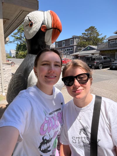 Two women are taking a selfie outdoors in front of a large puffin statue. The woman on the left is smiling and looking at the camera. She is wearing a white t-shirt with a graphic print. The woman on the right is wearing sunglasses and a white t-shirt with a Stüssy logo. She is also smiling at the camera. Behind them, there are buildings, trees, and a parked truck. The scene appears to be in a town with a coastal vibe, possibly Cannon Beach, Oregon, known for its puffin sculptures and beach town atmosphere. The sky is blue and the weather is sunny.