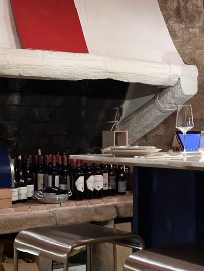 An interior image depicts a section of a commercial establishment, likely a restaurant or wine bar, in Caminha, Portugal. The background features a dark-toned stone or brick structure, possibly a large fireplace opening, topped by a thick, white-painted wooden beam. Above this beam, a white architectural element with a prominent diagonal red section is visible. A rough-hewn stone ledge directly below the beam displays approximately thirty to forty bottles of red wine, standing upright, with various labels and dark capsules. A silver-colored, oval serving dish with handles is placed among the wine bottles on this ledge, alongside a light-colored wooden box. In the mid-foreground, a light-toned countertop, supported by a dark blue base, holds a stack of white plates, silverware, and a single clear stemmed wine glass. An upright electronic tablet with a blue screen is positioned behind the wine glass. Two metallic, curved, backless bar stools are partially visible in the lower foreground. No individuals are present in the scene.