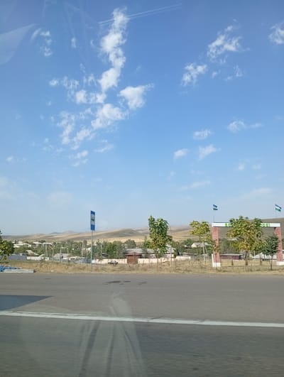 The image depicts a street scene in the city of Balgaly, Uzbekistan. In the foreground, a gray asphalt road is visible, with a white line running along its edge. Beyond the road, a strip of dry grass separates it from a village. The village consists of numerous houses with white and beige walls, some with fenced yards. Several trees with green foliage are scattered throughout the village and along its edge. A blue and white bus stop sign stands on a pole in the middle ground. Further back, a brick archway with white pillars is adorned with two flags. The archway has text written on it that appears to read "BALGALY" in white letters. The background features rolling, arid hills under a bright blue sky with scattered white clouds. The overall scene suggests a sunny day in a rural or semi-rural area of Uzbekistan.