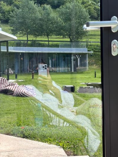 A building, a grassy lawn, and a tree are visible through a window. A modern, rectangular building with a glass facade is in the center of the frame. A manicured lawn surrounds the building, and a leafy green tree stands to the left. The reflection of a person holding a phone is visible in the glass, and a striped blanket is resting in the lower center of the frame. The window has a dark frame and a silver handle. The location is not identifiable as Chkalov, Armenia, based on the visual information.