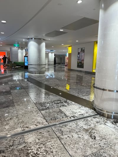 This media file captures an indoor view of a modern public space, likely a shopping mall or large commercial building in Indooroopilly, Australia. The perspective is angled, looking down a wide, well-lit corridor.

The setting is characterized by clean, contemporary architecture. The ceiling is white and curved, featuring numerous circular recessed light fixtures and rectangular ventilation grates. Several prominent, cylindrical columns punctuate the space. These columns are faced with small, light-colored rectangular tiles or textured panels and are adorned with metallic bands at the top and bottom.

The floor is a key visual element, composed of large, highly polished granite or marble tiles with a distinct speckled pattern of white, grey, and dark brown. A smooth, reflective grey strip runs longitudinally down the center of the visible floor area, creating a visual path. The high polish of the floor creates clear reflections of the overhead lights and distant elements, giving a sense of depth and spaciousness.

In the distance and along the sides, several retail displays or shopfronts are visible. One prominent display on a wall features a large image of a group of people, possibly an advertisement. Another shopfront in the foreground is brightly lit with an orange hue, where parts of the words "UP" and "YOU" are discernible on a sign. Bright yellow signs with partially legible text, such as "Say" or "Says," are also visible on the walls. A green exit sign is present in the middle ground.

A few indistinct human figures are scattered throughout the scene. Most appear as distant, dark silhouettes, casually walking or standing. One person in the foreground, dressed in dark clothing, is looking towards the orange-lit display. The overall activity suggests a typical, unhurried day in a public thoroughfare. No specific event or intense activity is taking place. The time of day is indeterminate due to the artificial indoor lighting.