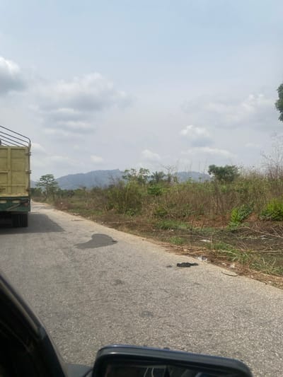 The image captures a daytime scene along a paved road in Sasaro, Nigeria, observed from the interior of a moving vehicle. In the left lane, the rear section of a light green flatbed truck, featuring high side railings, is visible. The asphalt road surface displays several dark, irregular stains. To the right of the road, a strip of sparse, dry vegetation, consisting of grasses and shrubs, extends towards a line of denser foliage and trees. A prominent range of hazy, bluish-grey mountains or hills forms the distant background beneath a sky with scattered clouds. A black rearview mirror from the observer's vehicle is partially visible in the lower right corner. No human figures or animals are discernibly present.