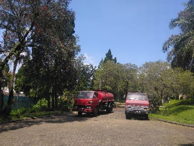 The image displays an outdoor scene in Nová Bystřice, Czechia, under a clear blue sky with bright sunlight. Two red utility vehicles are parked on a paved surface composed of dark grey interlocking pavers, which are sparsely covered with light-colored debris.
To the left, a red tank truck is parked, oriented diagonally towards the viewer's left. It features a large, horizontally mounted red cylindrical tank on its chassis. The front of the truck is visible, including the cab and front bumper.
To the right of the tank truck, an older model red van is parked, facing directly towards the viewer. Its front grille, headlights, and windshield are visible, though details like license plates are indistinct due to resolution.
The area is extensively vegetated. On the left, a large, dark-barked tree with dense green foliage and some scattered reddish blossoms or berries dominates the frame. Behind and between the vehicles, numerous other trees with green leaves form a dense backdrop. A bright green grassy slope with additional trees and shrubs borders the paved area on the right. In the far left background, a light-colored building is partially visible behind the trees, adjacent to a segment of a light green fence or barrier. A white horizontal barrier arm is partially visible behind the red van on the right. No individuals are present in the scene.