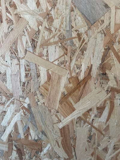 This media file is a close-up, full-frame photograph of oriented strand board (OSB). The image is an abstract composition of wood chips and strands of various sizes and colors, ranging from pale beige to dark brown and gray. The texture is rough and layered, with individual wood pieces overlapping and intermingling. Some strands appear smoother and more processed, while others show the natural grain of the wood. The lighting is even, indicating it was likely taken in a well-lit indoor setting or on an overcast day. There are no discernible subjects like people or animals, nor any specific objects or scene elements. The image captures the material itself, focusing on its granular and fibrous texture. The context provided indicates the OSB may be a material used in Orlivka, Ukraine, but the image itself offers no visual cues related to this specific location, time of day, or weather conditions. There is no visible text within the image.