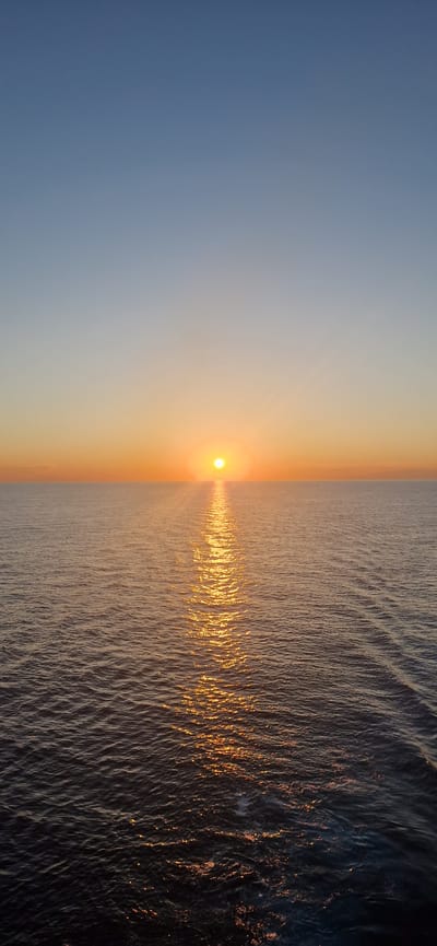 The image depicts a serene sunset over a calm body of water. The sun, a bright orb, is positioned low on the horizon, casting a golden path across the water's surface. The sky transitions from a deep blue at the top to a warm orange hue near the horizon. The water is a dark, reflective surface, with gentle ripples creating a textured pattern. The overall scene is tranquil and peaceful, with no visible signs of human activity or urban elements. The absence of any identifiable landmarks or structures makes it impossible to determine a specific location within the city of Monett, United States of America, or any other location for that matter.