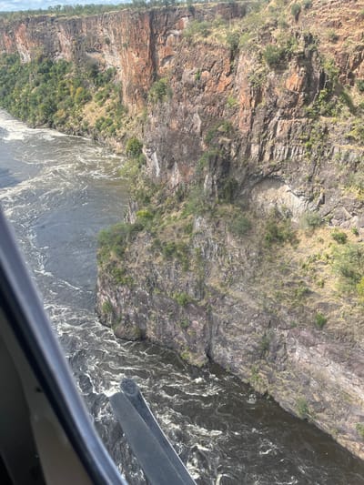 An aerial view captures the edge of a cliff overlooking a river in Victoria Falls Municipality, Zimbabwe. The cliff, displaying stratified rock formations, is sparsely vegetated with grass and shrubs. The river below appears turbulent with white water. The scene is viewed through what seems to be an airplane window.