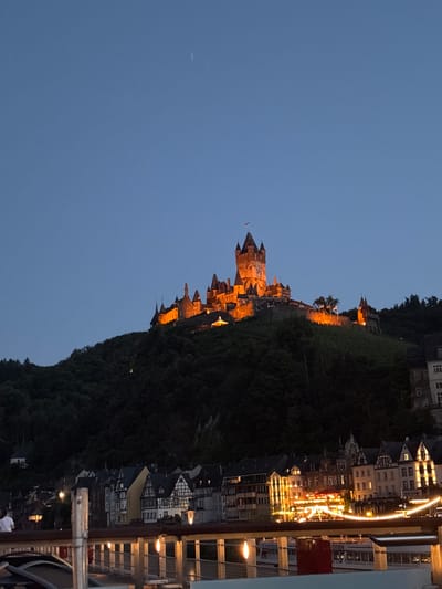 The image depicts a nighttime view of Cochem, Germany, with the Reichsburg Castle prominently illuminated on a hillside overlooking the town. The castle, a historic landmark, is the central subject, its turrets and walls glowing with warm orange light against the darkening blue sky. Below the castle, the town of Cochem is visible, with its buildings characterized by traditional German architecture, many featuring dark timber framing. Lights from windows and streetlamps illuminate the town. In the foreground, a railing with decorative lights suggests a vantage point from a waterfront promenade or bridge, with a boat visible in the lower right corner of the image. The environment is that of a serene evening setting in a European town.