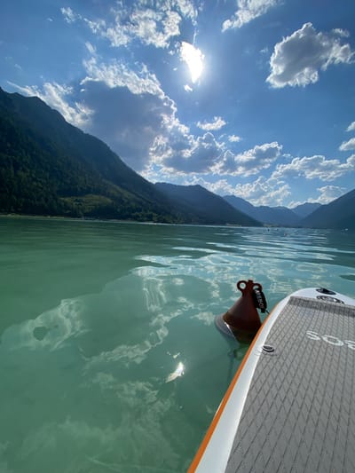 An over-the-shoulder, first-person perspective shot from a paddleboard on a serene, emerald-green lake in Lärchenwiese, Austria. The paddleboard in the foreground is gray with a diamond-patterned deck and an orange stripe. To the left of the board, a rusty-orange buoy with a black strap floats in the water. The lake's surface reflects the bright blue sky, scattered white clouds, and the sun, which is partially obscured by clouds in the upper right quadrant. Majestic, tree-covered mountains frame the lake in the background, their slopes descending towards the water. The water appears calm, with subtle ripples breaking the reflections. In the distance, several more buoys are visible, marking the expanse of the lake. The overall scene evokes a sense of tranquility and natural beauty.