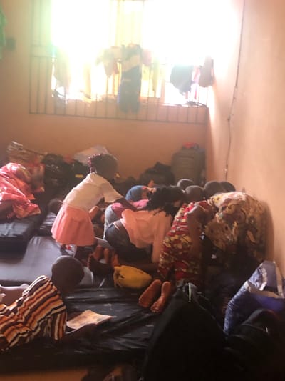 Several people, including children, are in a room in Nteje, Nigeria. The room has a barred window that is letting in bright light. The people are sitting on a bed or floor, surrounded by bags and clothing. Some are kneeling or bending over. One child wears a white and pink dress, and another wears a striped shirt.