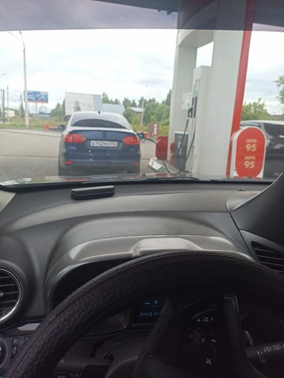 The image, taken from inside a car, shows a blue Volkswagen Jetta at a gas station. The car's license plate is visible and reads "B742MX 252". To the right of the Jetta are gas pumps, one of which displays "95" for gasoline prices. In the background, there are trees and buildings, suggesting an urban or suburban environment. The dashboard of the car is in the foreground, showing the steering wheel and instrument panel. The overall scene depicts a typical moment at a fuel station in Shakhunya, Russia.
