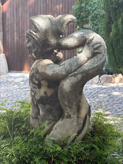 The image shows a stone gargoyle statue in an outdoor setting. The statue is weathered and features a horned, grotesque creature with its head bent down and its hands clutching its sides. The location appears to be a garden or courtyard, with stone paving in the background and lush green ferns growing around the base of the statue. A wooden fence or wall is visible behind the statue on the left. The specific location is Bunyola, Spain.