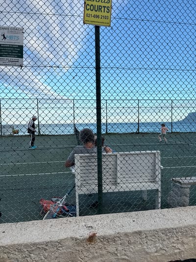 The image shows a tennis court in Muizenberg, South Africa, viewed through a chain-link fence with advertisements attached. The signage includes information for "Tennis Coach MBTS" with a phone number and website, and a yellow sign for "Tennis Courts" with a contact number. Two people are playing tennis on the court, one wearing black and white clothing and the other in red. Another person stands next to the court. A ball machine, rackets, and a bag are visible. In the background, beyond the tennis court, a body of water and the sky are visible. A concrete wall is present on the right side of the image.