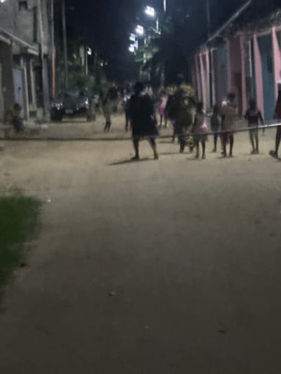 A blurry, grainy street scene at night. People walk along the sidewalk. Buildings line the left edge, with some lit by streetlights. The right side is mostly a gray-toned expanse, likely a road or open area, with some green vegetation at the bottom. The image has a general dark and unclear quality.