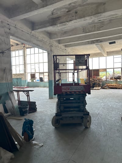 The image depicts the interior of an abandoned industrial building, likely a warehouse or factory, located in Djerman, Bulgaria. The space is characterized by concrete floors, columns, and a high ceiling with exposed beams and pipes. Large, multi-paned windows dominate the walls, allowing natural light to enter, though some are broken or boarded up. Debris, including piles of wooden pallets and construction materials, is scattered throughout the space.
A red scissor lift is positioned centrally in the foreground. It has "HAEM 0882252263 WWW.TOPMACHINE.COM" printed on its side. To the left of the scissor lift, a metal table with some items on it, a bucket, and building materials are visible. In the lower left corner, there are more construction materials, including wooden planks, and a blue tarp covering an object.
The scene suggests a state of disuse or ongoing renovation, with no people present in the image. The overall atmosphere is one of abandonment and industrial decay.