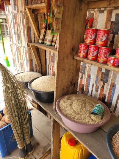 The image displays a detailed view inside a rustic market stall or small grocery store in Puerto López, Colombia. The background features a wall covered with patterned tiles or wallpaper depicting irregular rectangular shapes in brown, grey, white, and blue tones. Simple wooden shelves are affixed to this wall.
On the upper left shelf, various packaged goods, including several long, transparent bags of pasta or noodles with green and red branding, are visible. Below this, on a middle shelf, a grey plastic basin and a black plastic basin are filled with light-colored grains, consistent with rice.
To the right, on another section of the middle shelf, multiple red cylindrical cans are stacked. These cans are labeled "DE-ELO TOMATO PASTE" (or similar variant "TOMATO PATT"). Six cans are clearly visible in a staggered arrangement.
On the lower right shelf, a pink plastic basin is filled with the same light-colored granular material (rice), with a small green and white metal can partially embedded within it. Below this shelf, a bright yellow jerrycan with a red screw cap rests on a lower surface. A section of a black plastic basin, containing what appears to be light-colored beans, is partially visible in the bottom right corner.
In the left foreground, two traditional brooms made of dried plant fibers with dark bindings at their bases lean against a blue plastic crate. This crate contains several whole brown coconuts. No individuals are present in the scene.