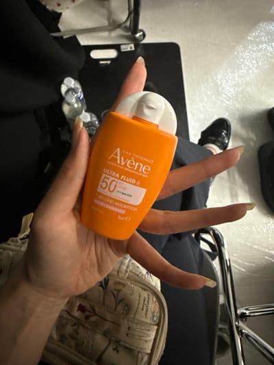 A fair-skinned hand, featuring long, natural-colored fingernails, holds an orange Avène Ultra Fluid SPF 50+ sunscreen bottle centrally in a high-angle perspective. The bottle's label displays "Avène EAU THERMALE LABORATOIRE DERMATOLOGIQUE ULTRA FLUID SPF 50+ UVB UVA BLUE LIGHT 1 SECOND ABSORPTION HAUTE RADIANCE 50ml e." Below the hand, a light-colored, zippered bag with a botanical pattern and partial text "Joanny A..." is visible. In the background, two clear plastic water bottles with white caps rest on a dark surface to the left. To the right, a dark blue fabric garment, a foot clad in a white sock and a black glossy shoe with a buckle, and a chrome-plated chair frame are discernible. The floor surface is light-colored, possibly concrete or tile, with a black mat situated beneath the chair. The specific location within Herentals, Belgium, is not ascertainable from the visual information provided.