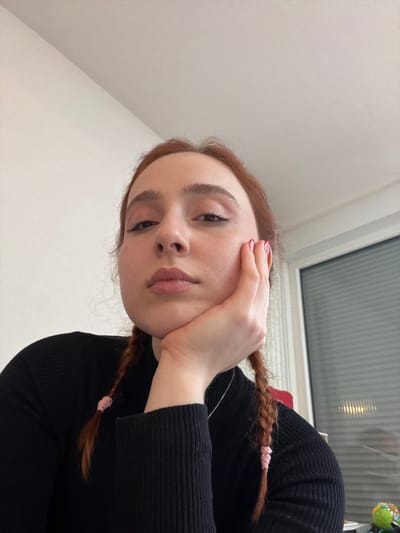 The scene is indoors, located in Eschau, France. A female individual is depicted in a close-up, high-angle shot. She has reddish-brown hair styled in two distinct pigtails, each secured with a pink elastic band. Her gaze is directed upwards towards the camera. Her chin rests on her right hand, which has fingernails painted pink with white patterns. She wears a black ribbed turtleneck garment and a thin silver necklace. The background consists of a plain light-colored wall on the left side of the frame. To the right, a window with horizontal blinds is visible, reflecting some light. A small, partially visible object, possibly a toy or decorative item with green, yellow, and blue colors, is present in the lower right corner, resting on an unseen surface.