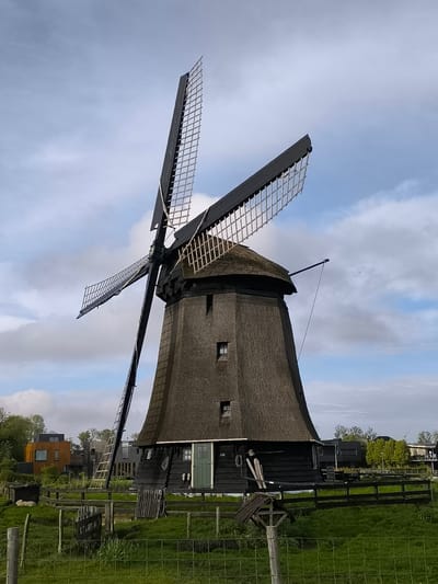 The image depicts a historic windmill in Oudorp, Netherlands. The windmill has a conical thatched roof and a black-painted wooden body. Four sails with lattice-like construction extend from the top. A ladder leans against one side. The mill is surrounded by a wooden fence and green grass. In the background, residential buildings can be seen. The sky is filled with clouds.