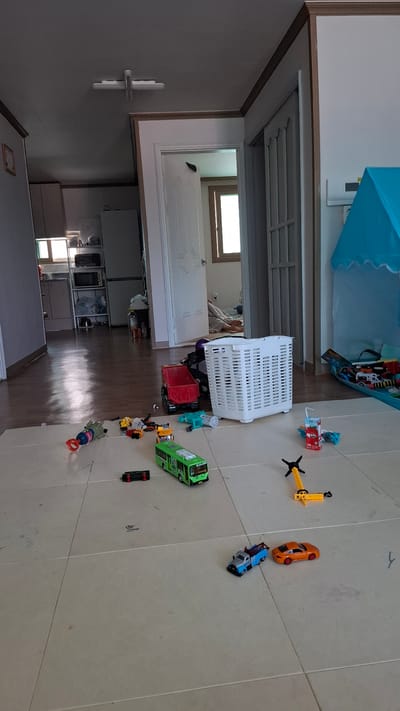 The image depicts a cluttered indoor scene, likely a child's play area within a residential dwelling in Daeso, South Korea. The floor is tiled and strewn with various toys, including several toy vehicles: a green bus, a red dump truck, a tow truck, and an orange sports car. A white plastic laundry basket filled with items is centrally located, and a blue play tent is visible to the right. In the background, doorways lead to other rooms, revealing glimpses of furniture, appliances, and a person lying on the floor. The lighting suggests it is daytime.