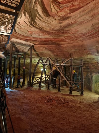 The image shows the interior of the sandstone caves in Homburg, Germany. The caves have layered sandstone walls in hues of red and brown. A metal structure with multiple levels spans the space, and two levels contain people. These individuals are dressed in dark clothing and appear to be wearing yellow hard hats. The metal structure is composed of a cage-like framework with crossbeams, platforms, and what appears to be netting. The caves are lit by artificial lights, casting shadows and illuminating the rocky texture of the sandstone. Cables and pipes are also visible, running along the lower portion of the structure.
