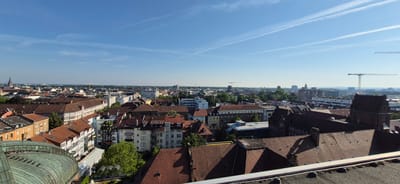 The image is an aerial view of a city, likely taken from a high vantage point such as a tower or tall building. The sky is blue with thin white contrails from airplanes. The city below is densely packed with buildings of various architectural styles and heights, many with red tile roofs. In the foreground, part of a green domed structure is visible on the left. In the midground and background, there are numerous residential and commercial buildings, some with balconies and windows. Trees are interspersed among the buildings, indicating green spaces within the urban environment. Several construction cranes are visible in the distance, suggesting ongoing development. The city appears to be European in character, with the architecture suggesting a location in Germany. Specifically, the architecture and city planning are consistent with the city of Karlsruhe, Germany, potentially viewed from the vicinity of the Karlsruhe Castle.