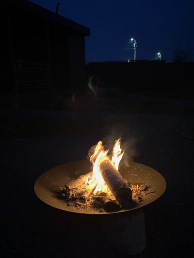 The image depicts a fire pit with burning logs in the foreground, set against a dark, twilight sky. A portion of a building with horizontal siding is visible on the left, silhouetted against the sky. In the background, two streetlights on poles illuminate the scene. The ground around the fire pit appears to be covered in gravel. The overall atmosphere is serene and dark. The location is identified as Великие Озера Кп, Russia.