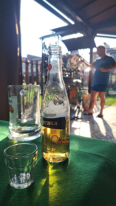 The scene depicts a partially consumed bottle of Baltika lager, accompanied by two drinking glasses on a green surface, possibly a table covering. The bottle label indicates the drink is brewed in Saint Petersburg, Russia. The image appears to have been taken outdoors, with a blurred figure visible in the background, moving from left to right. Structural elements like a roof and fence are also visible in the background. The location is identified as Рыбхоз, Russia.