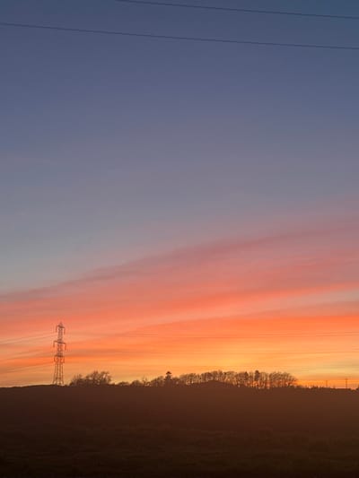 The scene depicts an outdoor rural landscape at either sunrise or sunset in Fowlis Easter, United Kingdom. The sky, occupying the upper two-thirds of the image, displays a gradient of colors transitioning from a muted blue-grey at the top to vibrant orange and pink hues with horizontal cloud streaks closer to the horizon. A large, metallic lattice-work electricity pylon is prominently positioned on the left side, silhouetted against the bright sky. Multiple dark power lines extend horizontally from the pylon and across the frame. A dark, silhouetted tree line stretches across the horizon, visible below the vibrant sky. The immediate foreground consists of a dark, indistinct landmass, likely a field, in shadow. No individuals, animals, or discernible actions are present.